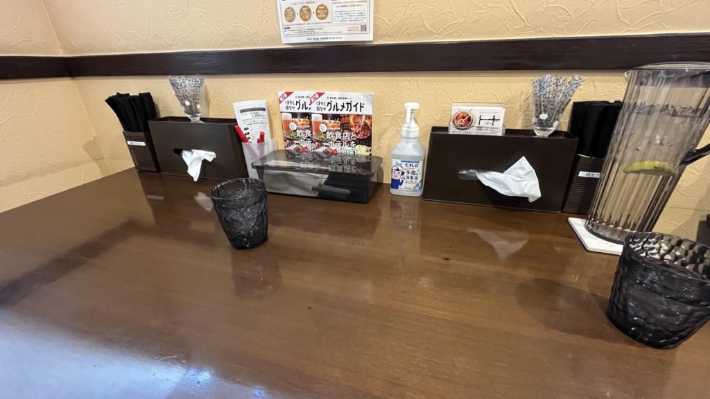 席の様子。茶色のテーブルの奥に、紙エプロン、ティッシュ、スプーン入れ、消毒液、水のピッチャーなどが置かれており、十分なスペースもある。